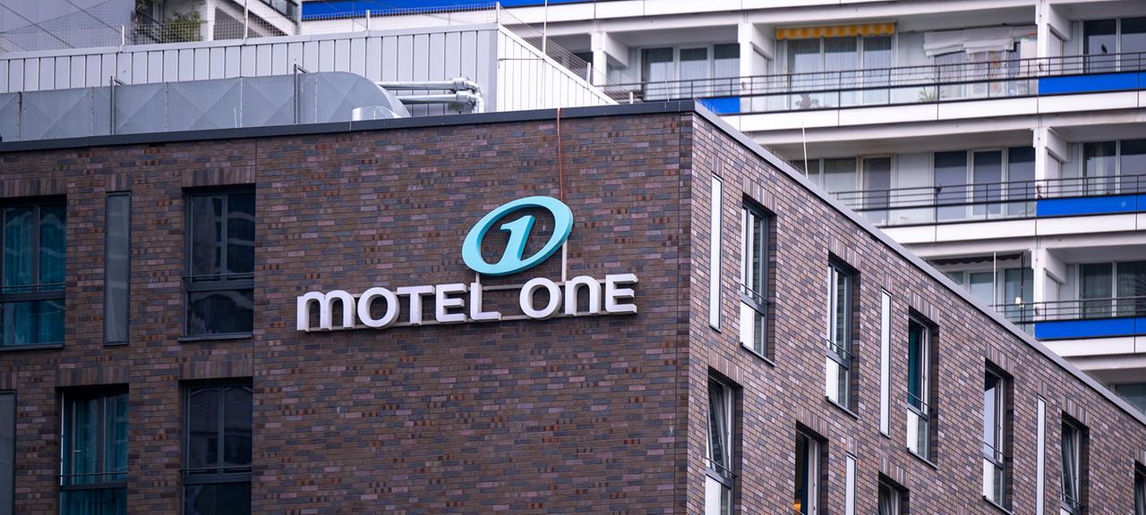 Eine Leuchtreklame mit dem Logo Hotelkette Motel One ist an der Fassade eines Hotels angebracht.